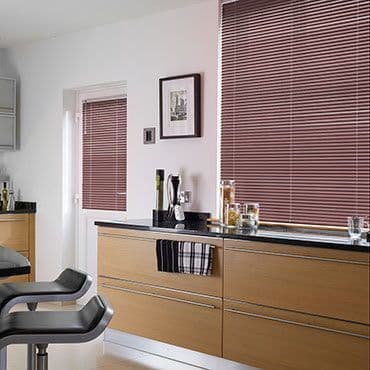 Brown horizontal slatted blinds cover a large window and a smaller door window, lowered and filtering daylight; modern kitchen with light wood cabinets, black countertop, bar stools, and glass jars.