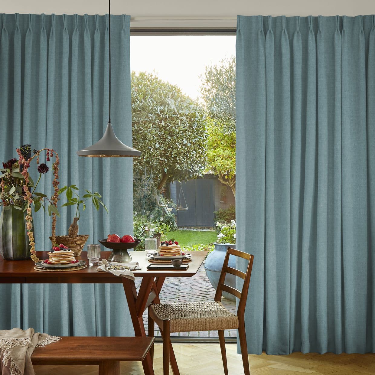 Teal pleated floorâ€‘length curtains, drawn aside to frame a central sliding door; filtering soft daylight. Dining table with stacked pancakes and pendant lamp in dining area overlooking a leafy backyard.