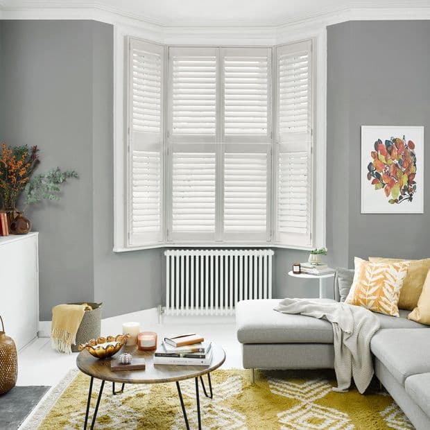 White plantation shutters covering a three-panel bay window, louvers tilted closed to diffuse soft daylight into a grey living room with radiator, sectional, yellow cushions, round coffee table and rug.