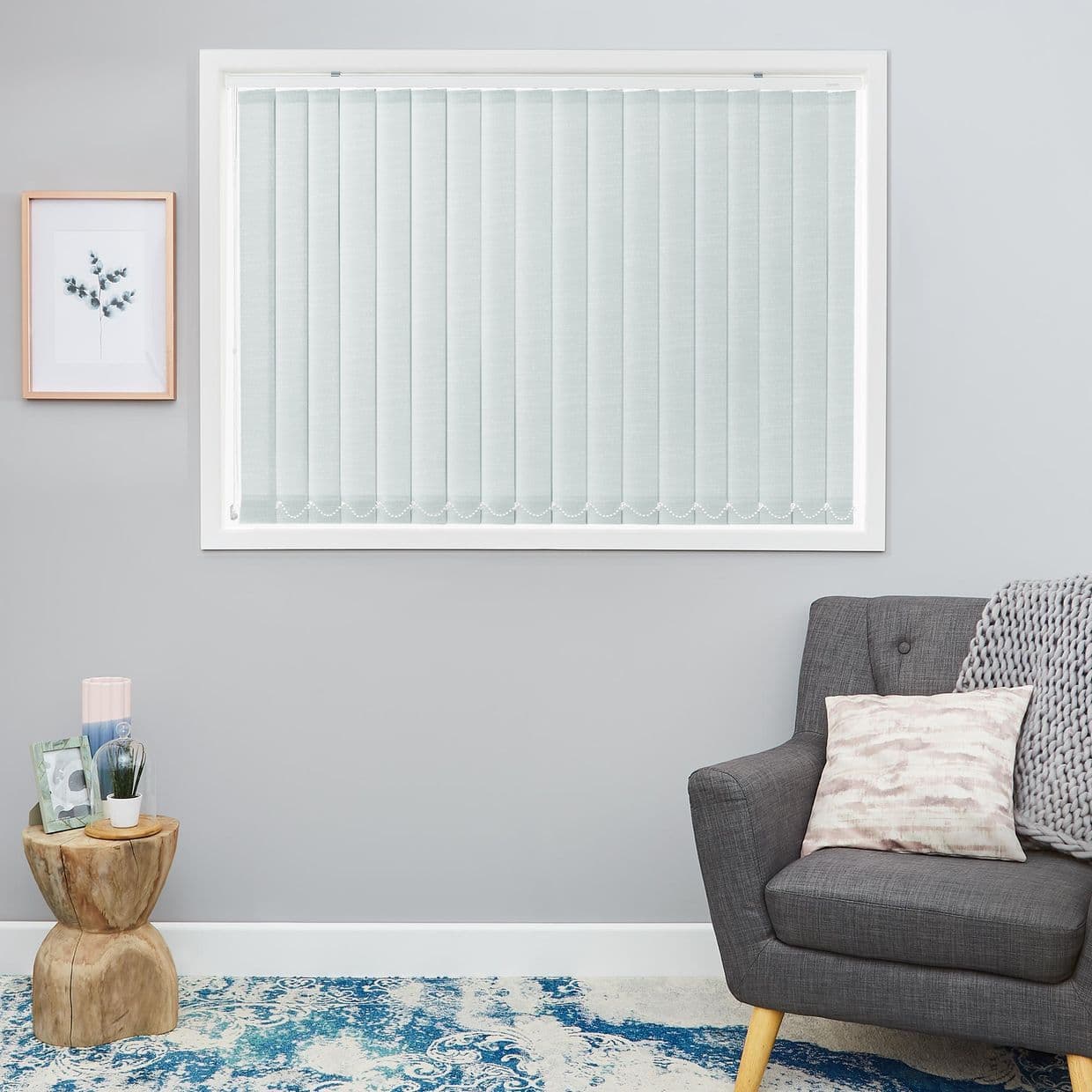 Vertical fabric blinds across a central window, closed and aligned with beaded weights, filtering soft daylight into a pale-gray living room with gray armchair, knit throw, wooden side table.