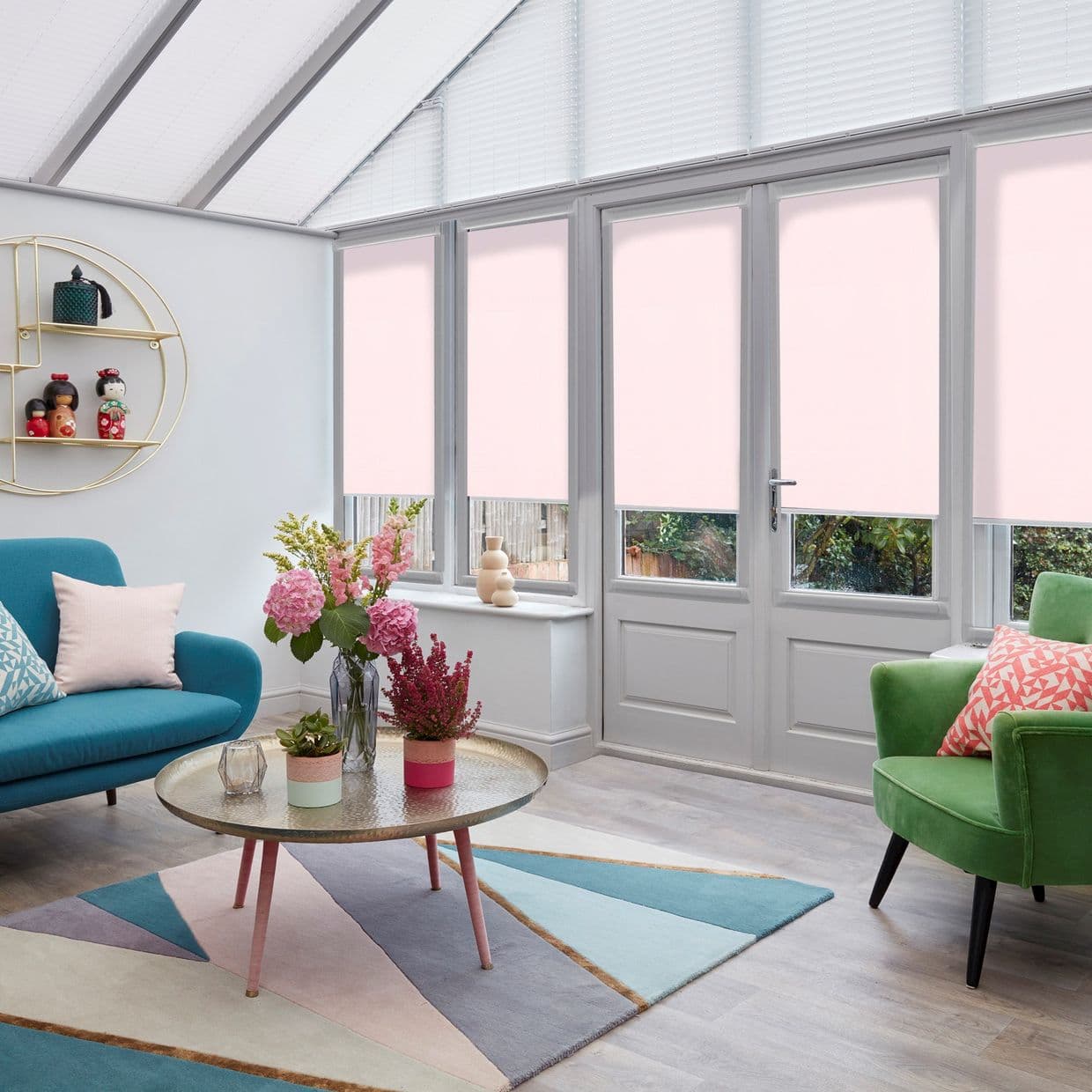 Roller blinds in pale pink cover tall French windows; pleated white blinds fill triangular roof panes, partially lowered to soften light in a bright conservatory with colorful seating.