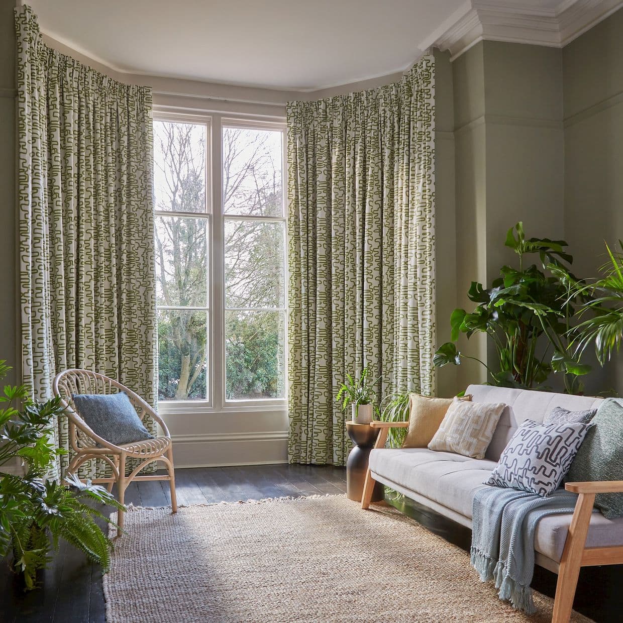 Green-and-white patterned, floor-length pleated curtains framing a large bay window, partially drawn and filtering soft daylight, in a cozy living room with pale-green walls, rattan chair, sofa and plants.