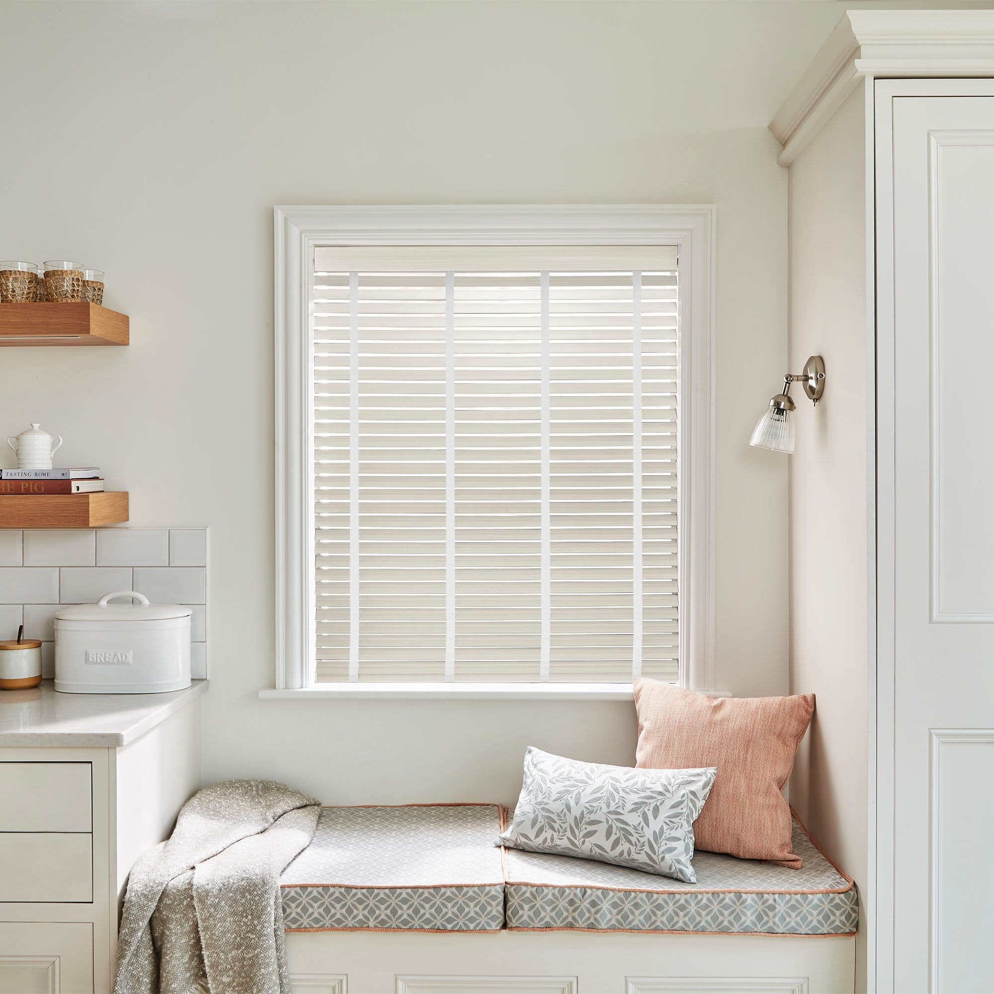 White horizontal venetian blinds covering the window, fully lowered with narrow slats filtering soft daylight; kitchen nook with built-in bench, patterned cushions, throw, wooden shelves, bread box, sconce.

BREAD
