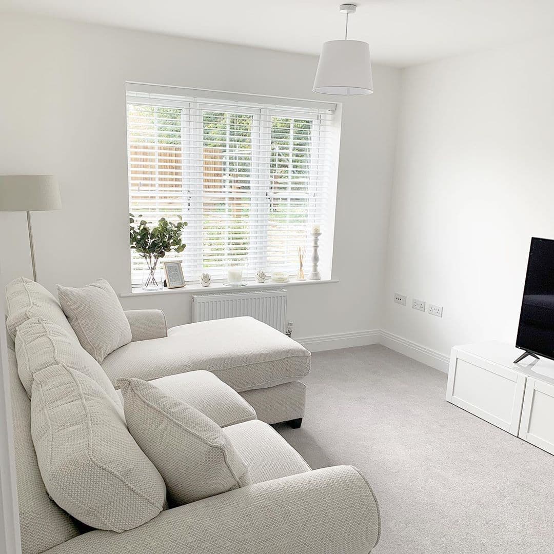 White horizontal wooden blinds covering a recessed window, slats tilted to admit diffused daylight; in a minimalist, all-white living room with pale upholstered chaise sofa, TV and carpet, garden outside.