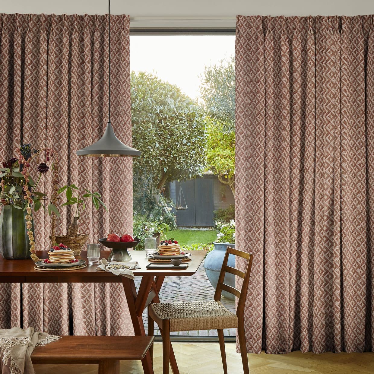 Pleated geometric rose curtains, parted at center and filtering soft daylight; in a breakfast dining area with pancakes and a pendant lamp, opening onto a leafy backyard garden.
