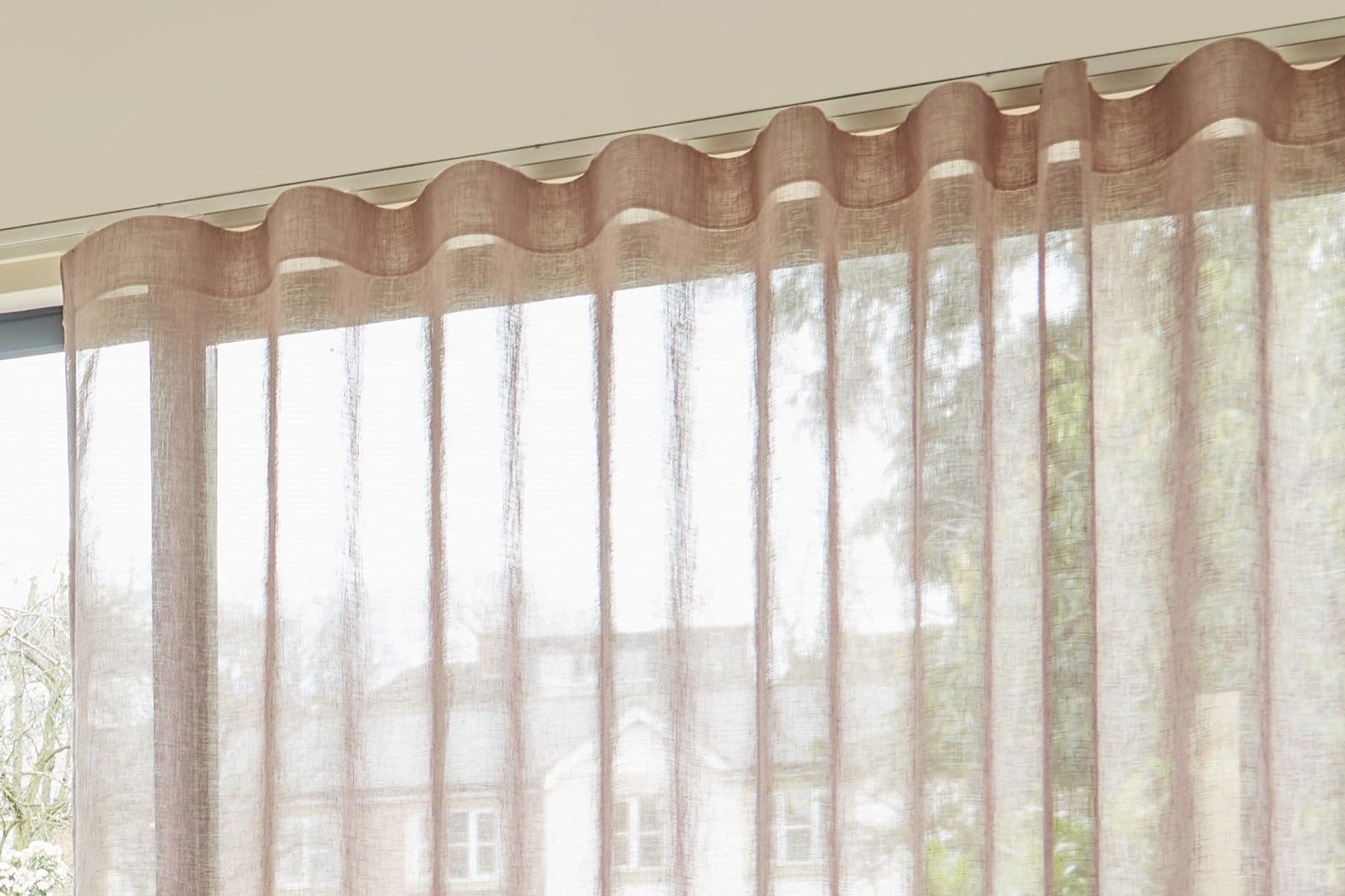 Sheer beige wave-headed curtains (ripple-fold) â€” hanging full-length, forming soft vertical pleats â€” filtering bright daylight into an interior living space, partially revealing blurred houses and trees outside.