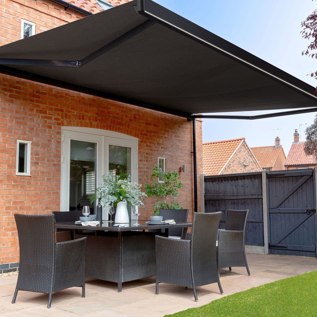 Large black retractable awning mounted above patio doors, fully extended with metal arms and LED trim; shading a wicker dining set on a brick-walled backyard patio under clear daylight.