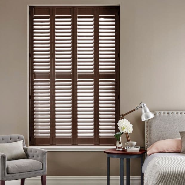 Dark wooden plantation shutters, four tall panels with adjustable louvers, closed and filtering daylight; set within a recessed window in a neutral-toned bedroom beside an upholstered bed and armchair.