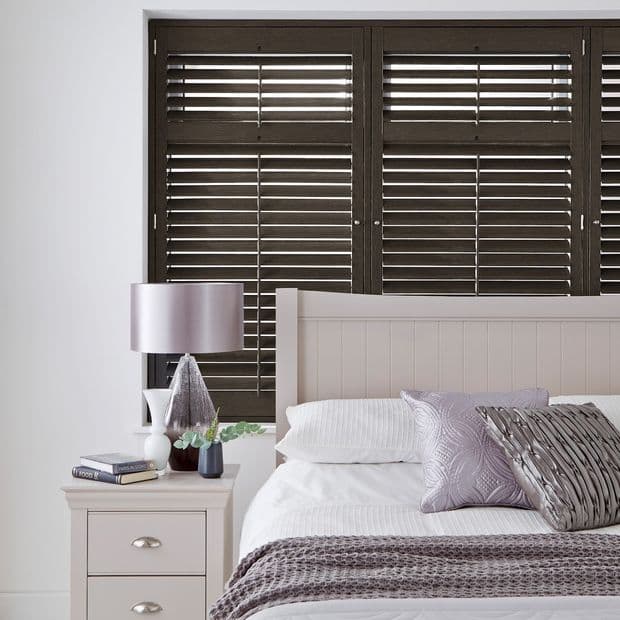 Dark wooden horizontal slatted shutters spanning the window behind the bed, slats partially tilted to filter thin light; bedroom setting with pale headboard, nightstand, lamp, pillows and soft lavender accents.