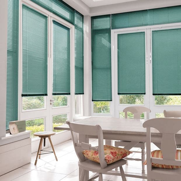 Teal horizontal Venetian blinds covering tall paneled windows and glass doors, partially lowered and filtering soft daylight. Context: bright sunlit dining nook with white table, chairs, and garden view.