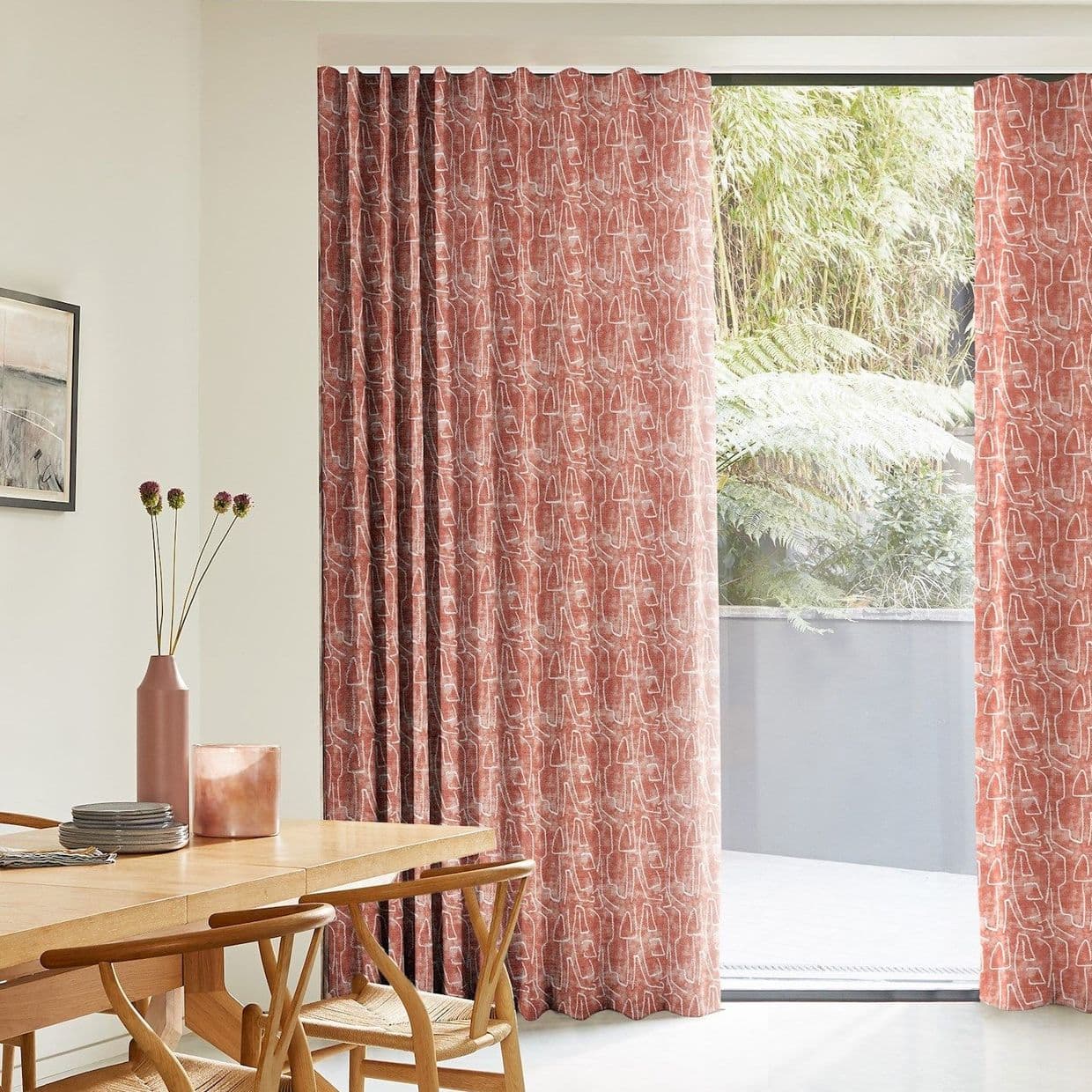Patterned rust-colored full-length curtains with an abstract white motif, partly drawn across a sliding glass door, filtering soft daylight into a minimalist dining area with wooden table, chairs, and greenery outside.