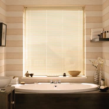 Cream horizontal Venetian blinds covering a bathroom window, fully lowered and closed, diffusing warm daylight onto an oval bathtub by striped beige walls with shelves, towels and decorative items.