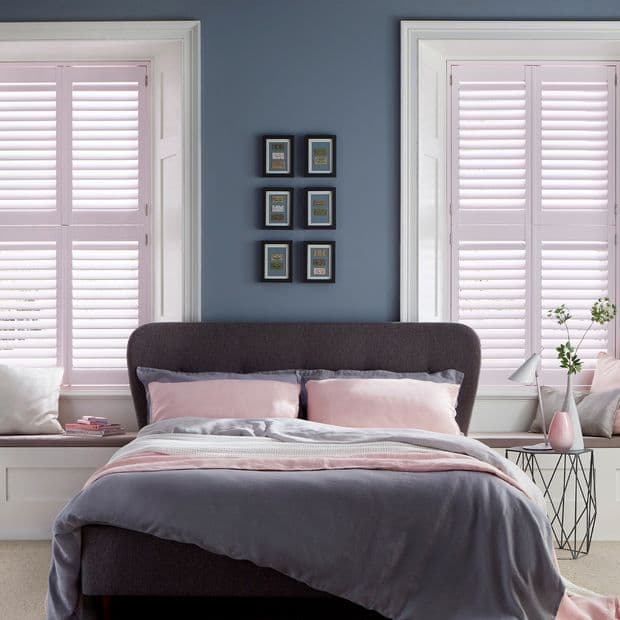 White interior plantation shutters on two flanking windows, closed with horizontal slats filtering soft daylight; a gray upholstered bed with pink bedding centered against a blue wall in a bedroom.