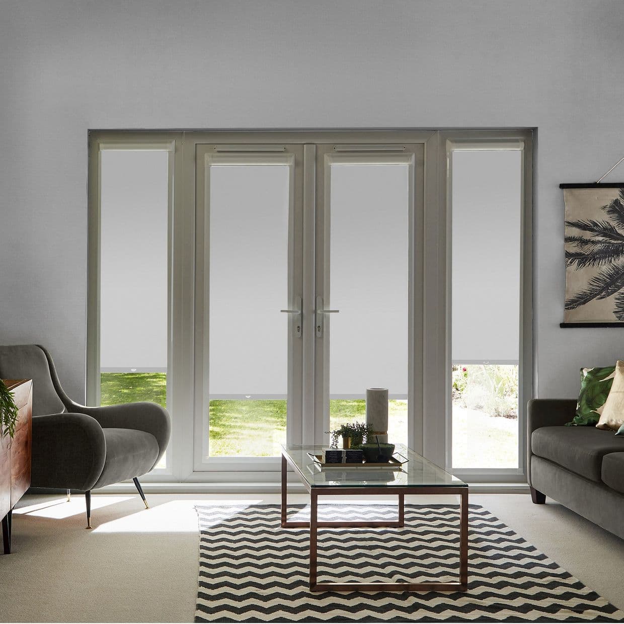 Roller blinds across four tall window panels, lowered three-quarters, light-gray, blocking most view but leaving narrow lower strips; living room with daylight, sofas and glass coffee table.