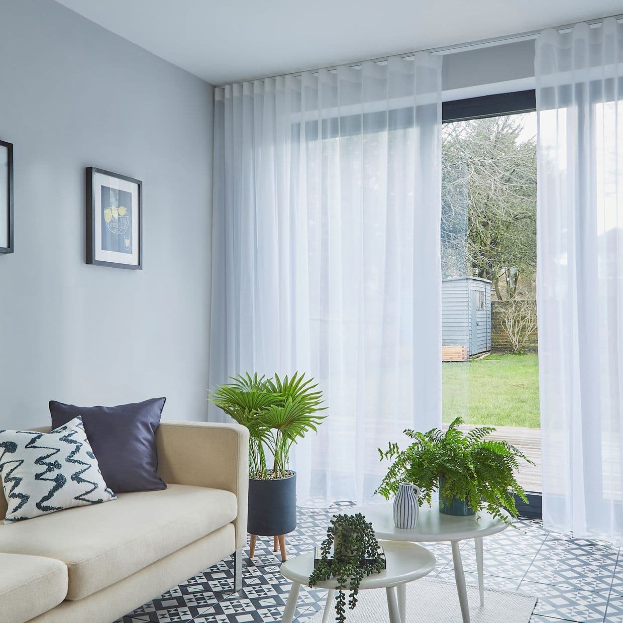 Sheer white floor-to-ceiling curtains on a ceiling track, filtering daylight through sliding glass doors; bright living room with beige sofa, patterned tile floor, plants, garden and shed view.