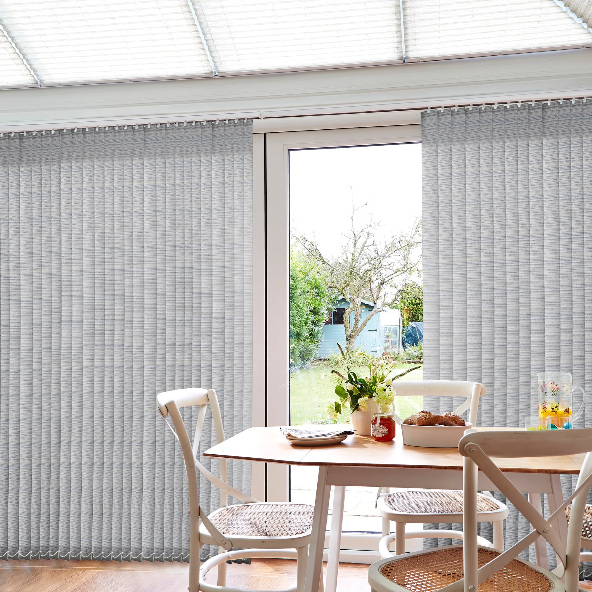 Vertical fabric vertical blinds covering sliding glass doors, partially drawn to reveal a central glass panel; softly diffusing daylight in a bright dining nook with wooden table and garden view.