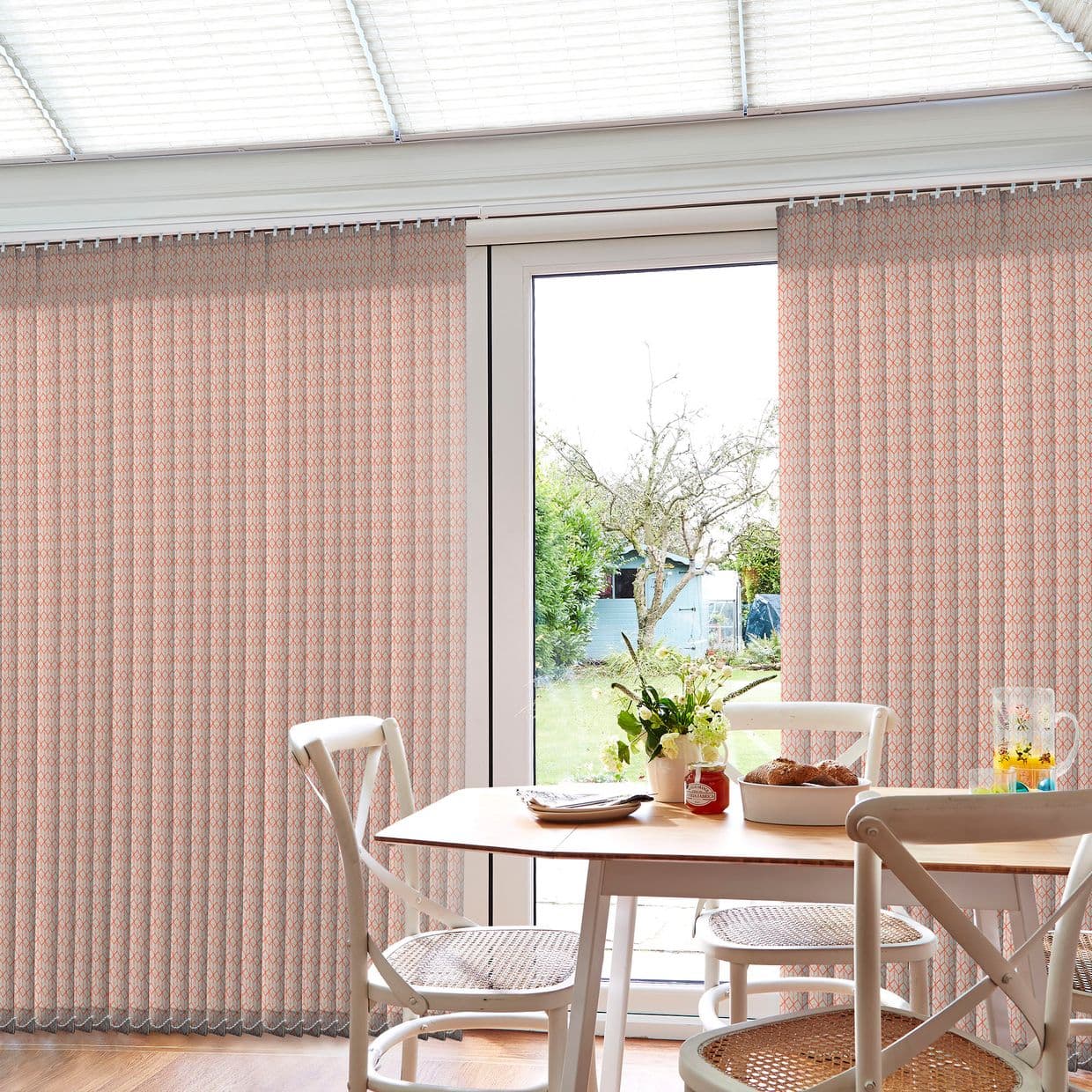 Patterned vertical curtains (pale pink geometric print) hang full-length, parted at the center to reveal glass door; filtering daylight over a dining table with cane-seat chairs and garden view.
