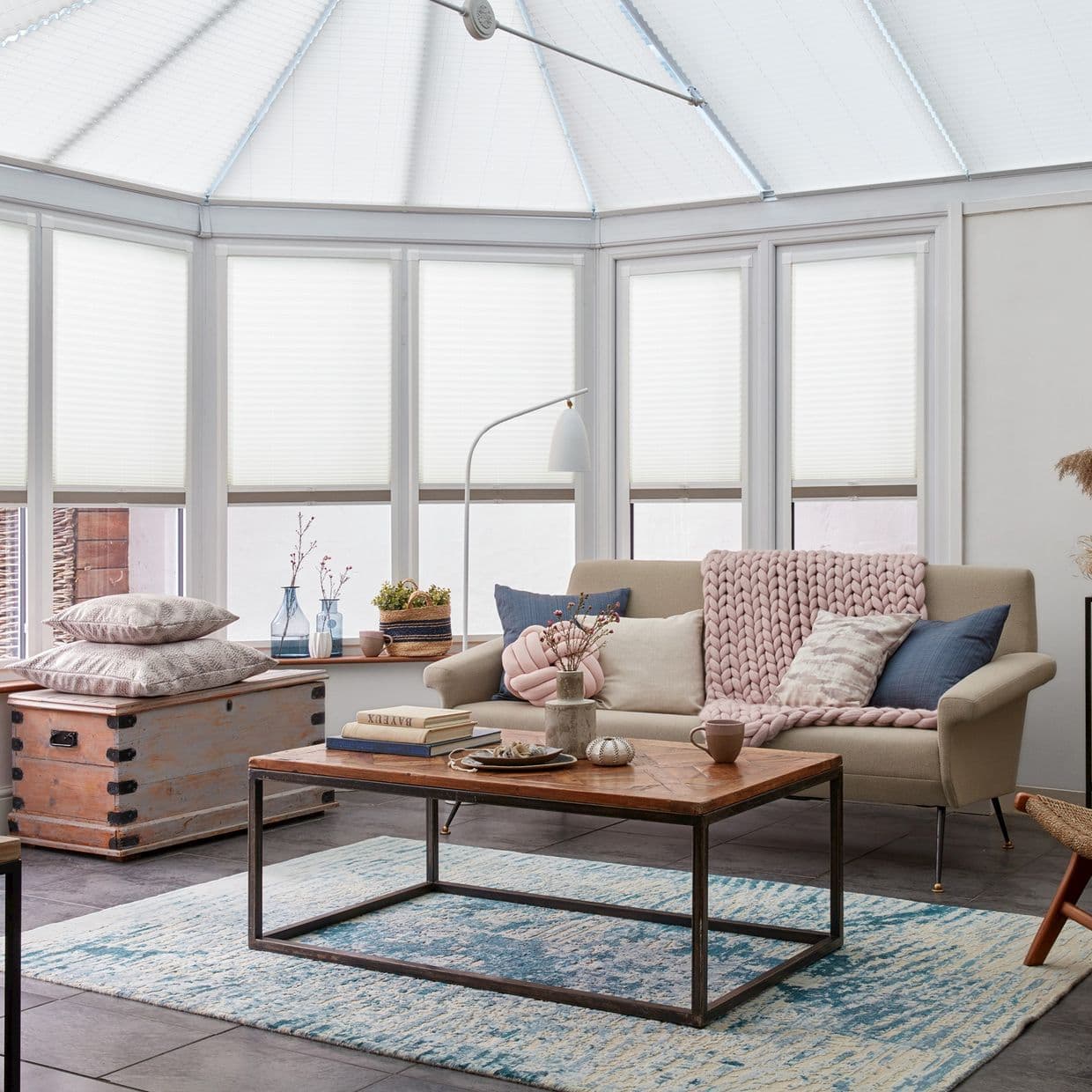 Cellular pleated blinds covering tall bay windows and conservatory roof, lowered to diffuse daylight; cozy sunroom living area with beige sofa, wooden coffee table, pastel throw and blue rug.