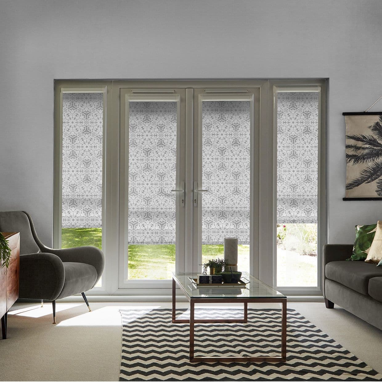 Patterned roller blinds on French doors and sidelights, lowered to diffuse light and obscure the view with narrow lower gaps showing lawn; contemporary living room with armchairs, glass coffee table, chevron rug.