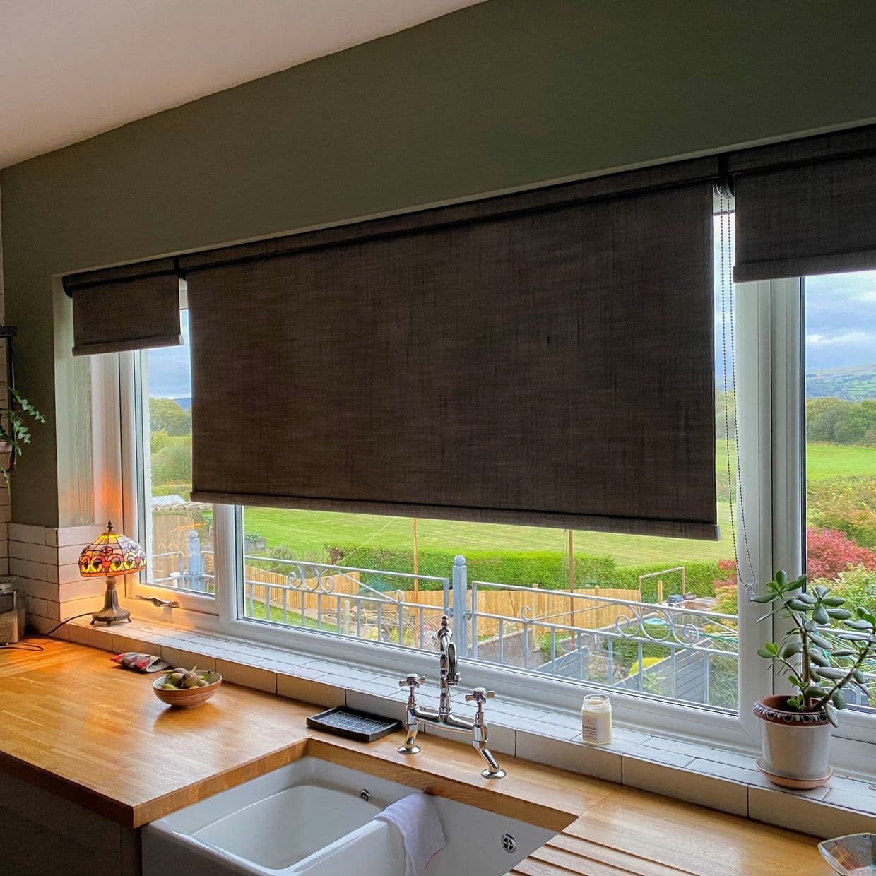 Dark grey fabric roller blind lowered across a wide kitchen window, partially filtering daylight; fitted pull-chain visible. Wooden countertop with sink and plants, warm lamp glow, green field view outside.