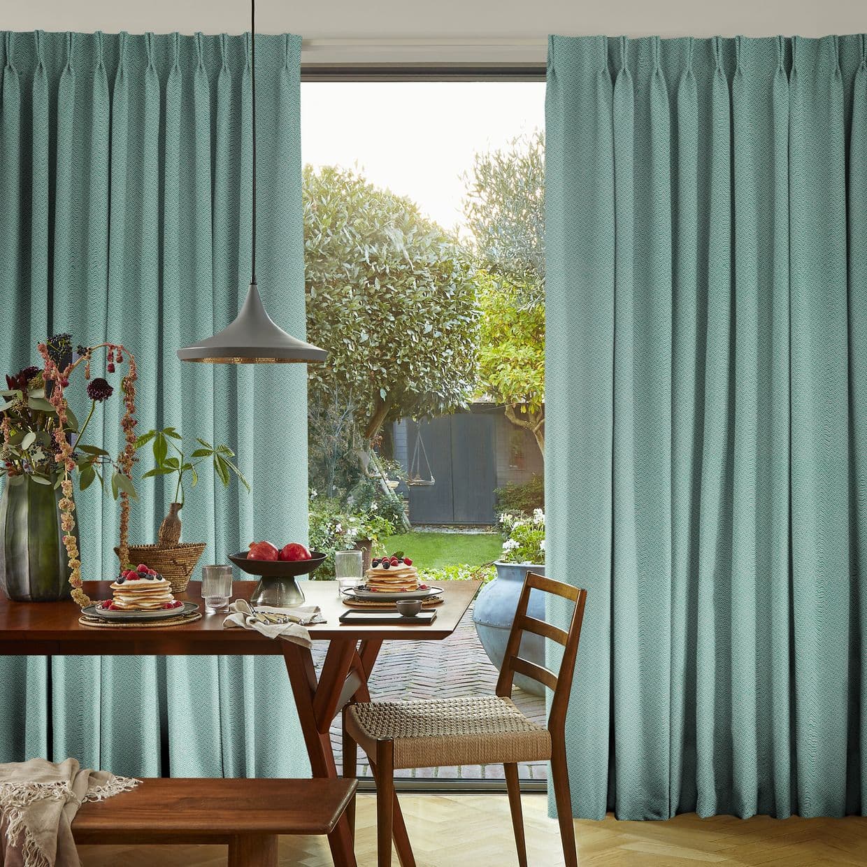 Teal pleated, floor-length curtains, parted at center to reveal a sunlit garden, framing a dining table set for breakfast with a pendant lamp, wooden chairs, and greenery outside.