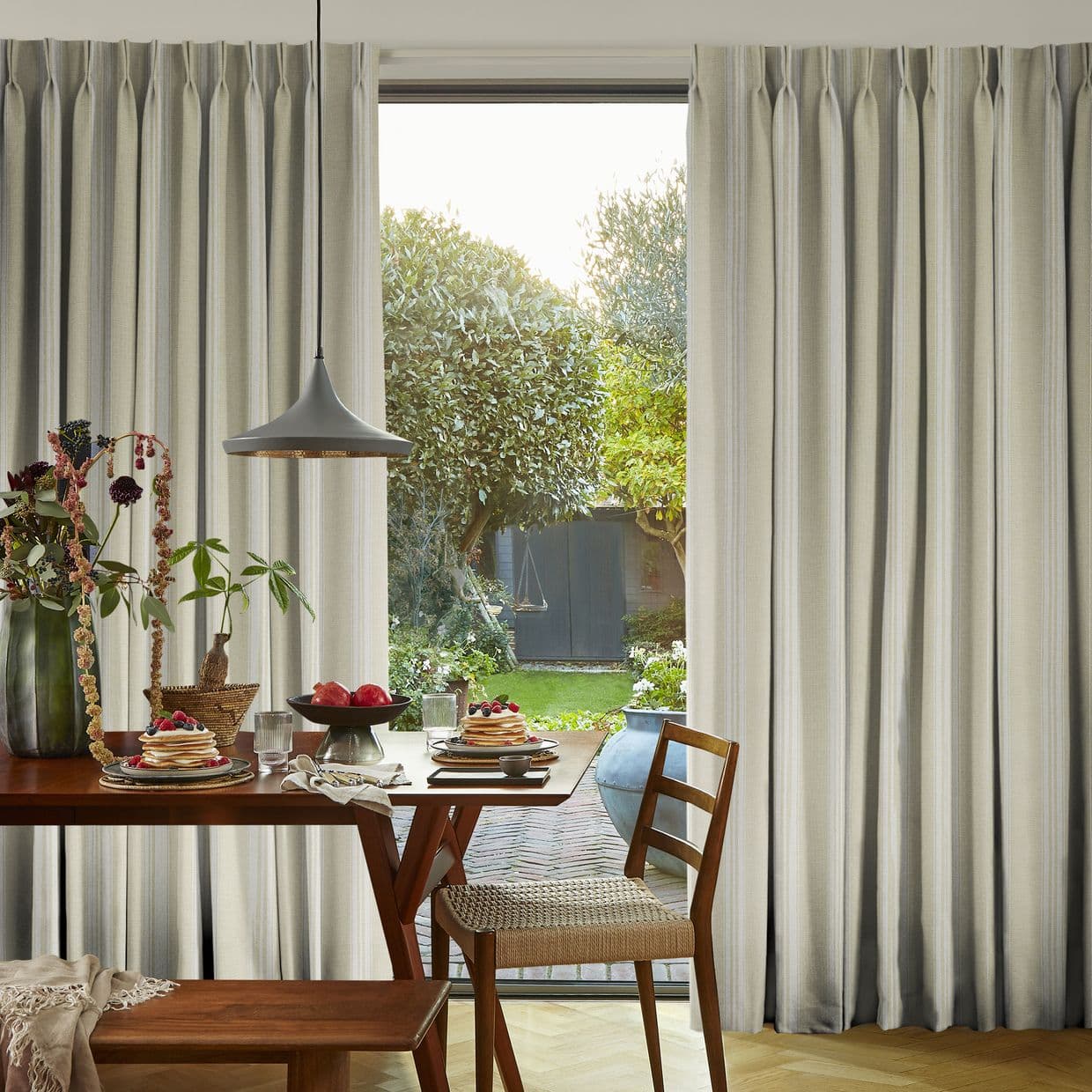 Floor-to-ceiling beige pleated curtains, parted centrally to expose glass door, softly filtering daylight; framing a dining table with pendant light, plates of pancakes, a chair, and a sunlit garden view.
