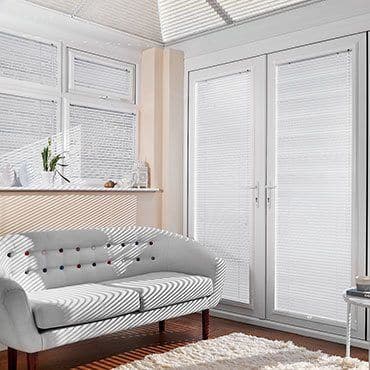 White pleated blinds covering upper windows and French doors, closed and filtering sunlight into striped shadows across a tufted light-gray sofa in a bright living room with hardwood floor.
