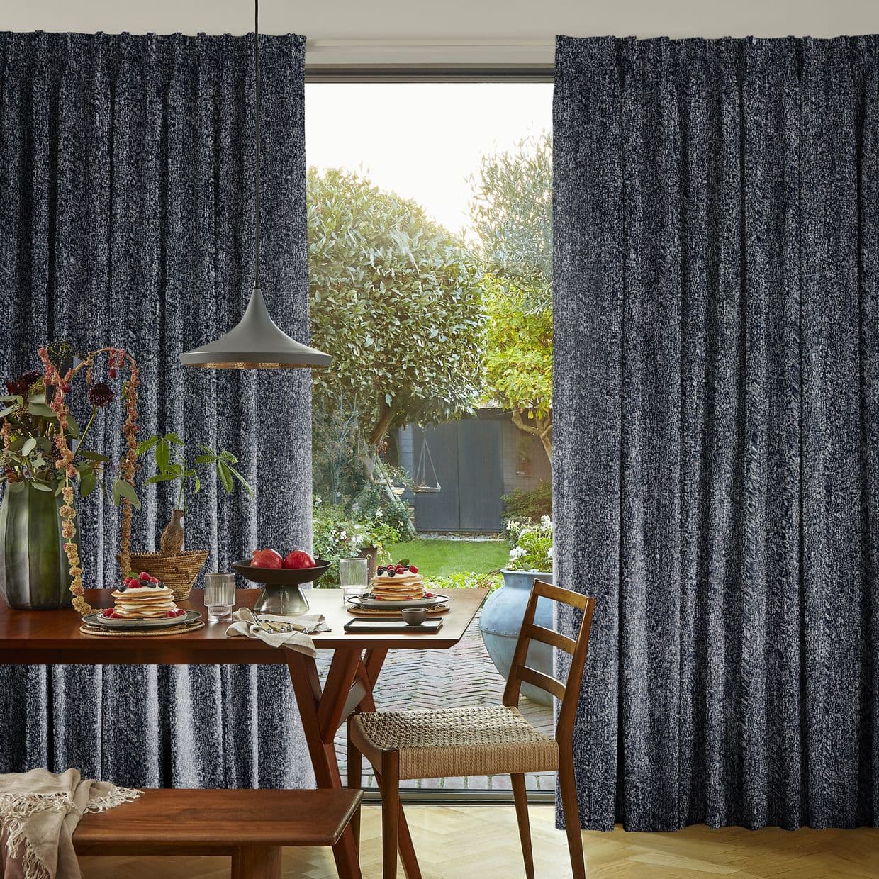Dark gray textured pleated floor-length curtains, drawn aside at center to frame the opening; open, admitting bright daylight and a lush backyard view beyond a dining table.