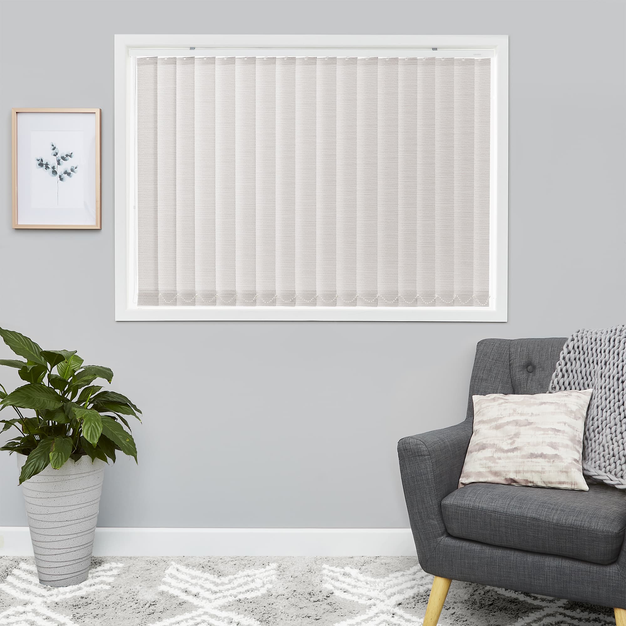 Vertical fabric blinds, beige and fully closed, cover a white-framed window, softly diffusing daylight; set in a gray living room with potted plant, framed botanical print, patterned rug and armchair.