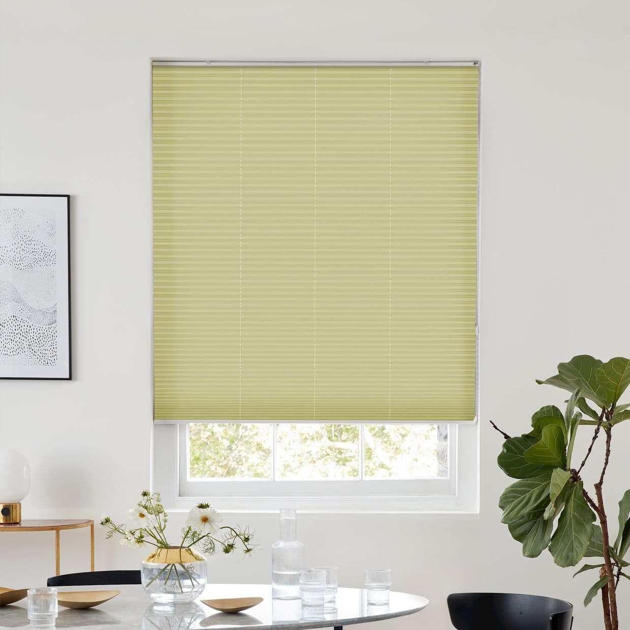 Light green pleated cellular blind, fully lowered across the window, filtering soft daylight; bright dining nook with white walls, round table with vase and glassware, framed art and a potted fig.