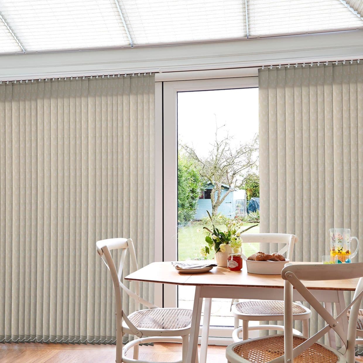Beige vertical blinds spanning a patio door, partially drawn to reveal the central glass door; filtering bright daylight into a small dining nook with wooden table, chairs, and backyard view.