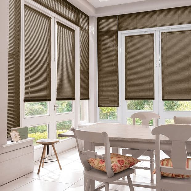 Brown horizontal Venetian blinds covering bay windows and glass doors, lowered and partially filtering daylight; bright dining nook with white table and chairs, floral cushions, tiled floor and garden view.