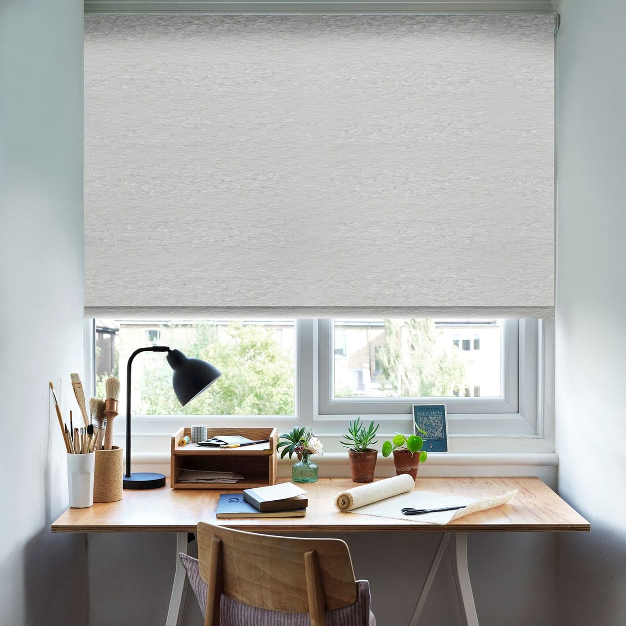 Light-grey textured roller blind, lowered with control at right, filtering soft daylight; a tidy home desk beneath with lamp, potted plants and notebooks, view of trees and nearby houses.