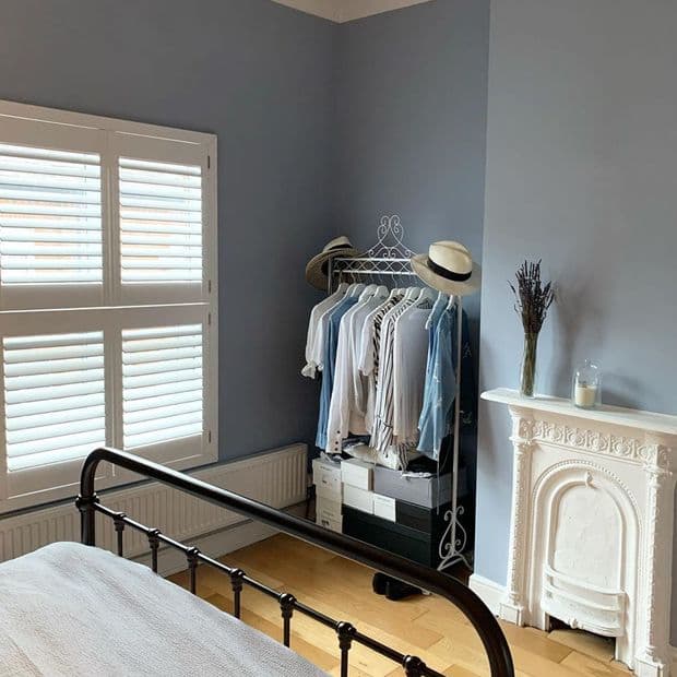 White plantation shutters on a window, closed with angled slats filtering soft daylight; blue-gray bedroom with black metal bedframe, open clothing rack of shirts and hats, hardwood floor, decorative mantel.