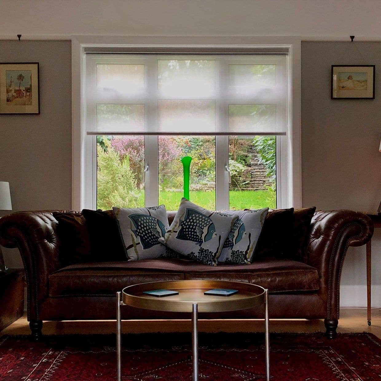 Translucent white roller blind, lowered halfway and filtering daylight; in a living room behind a brown leather sofa with patterned cushions; view of lush garden and stone wall.