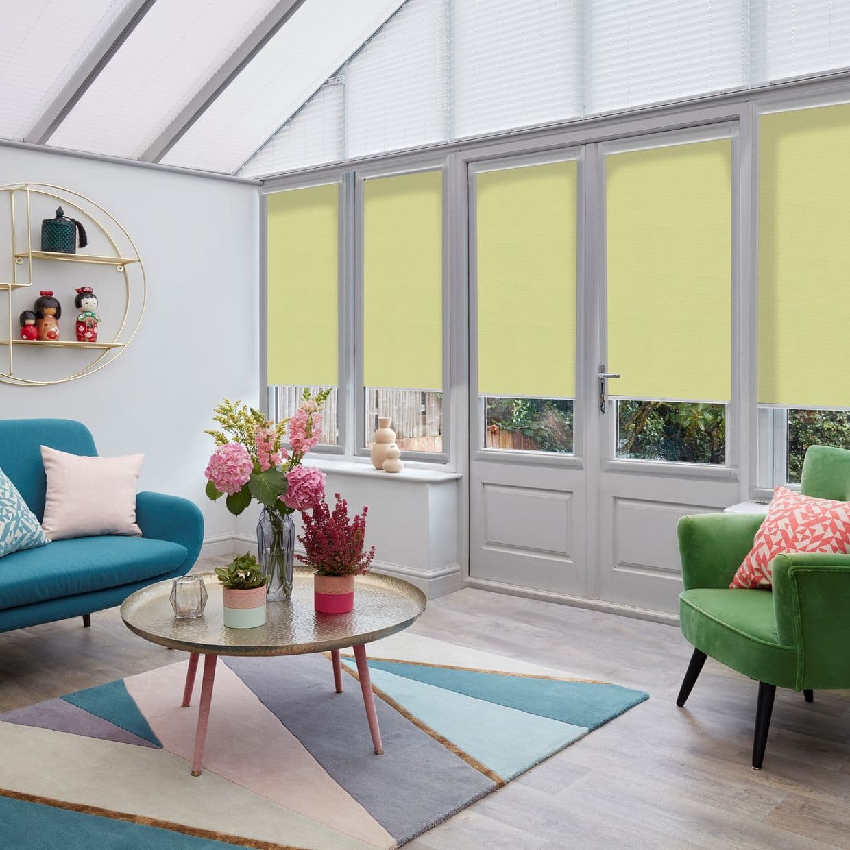 Yellow roller blinds, lowered covering most of tall patio windows, filtering daylight; in a bright conservatory living room with teal sofa, green armchair, round coffee table and flowers.