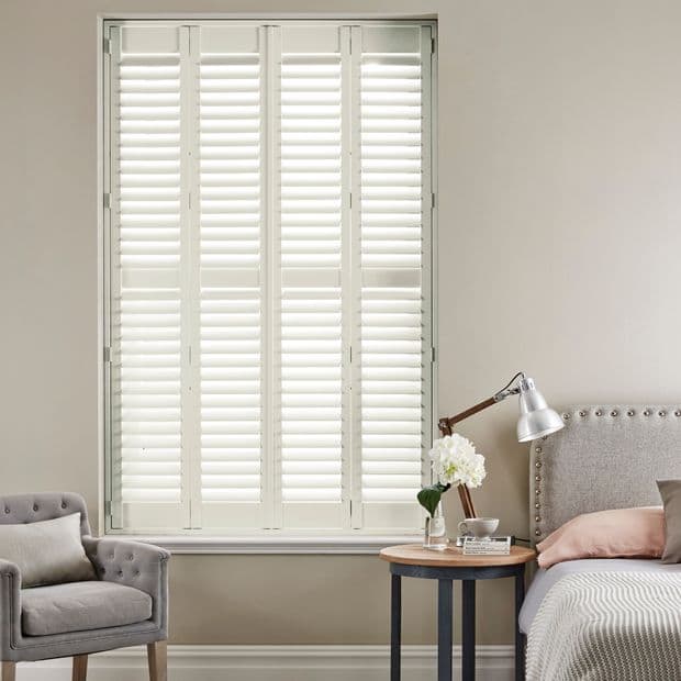 White plantation shutters covering a tall window, closed with angled louvers filtering soft daylight; bedroom setting with upholstered bed, round side table with lamp and flowers, armchair, and neutral walls.