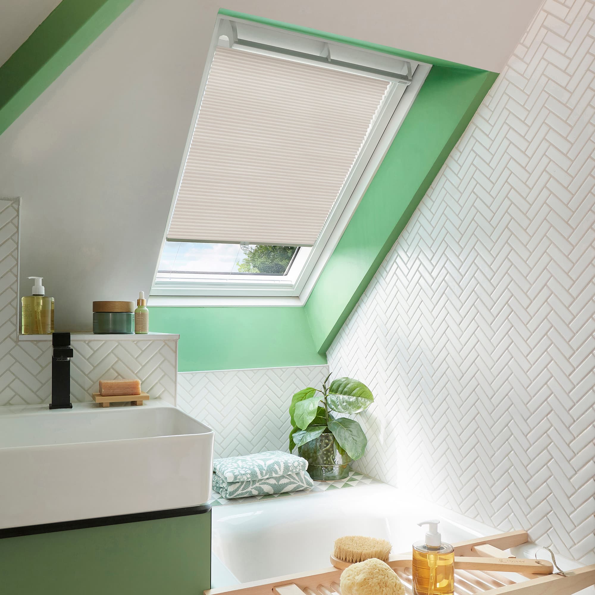 Cream pleated cellular blind lowered over a skylight, filtering daylight; set in a bright attic bathroom with mint-green window frame, white herringbone tiles, bathtub, sink, plant and toiletries.
