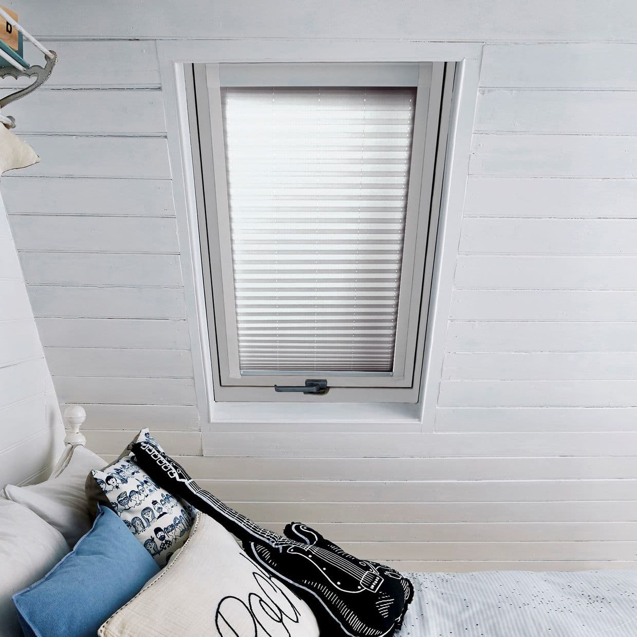 White pleated blind on a skylight windowâ€”closed, filtering soft daylightâ€”set in painted tongue-and-groove attic ceiling above a bed with pillows and a black guitar-shaped cushion.