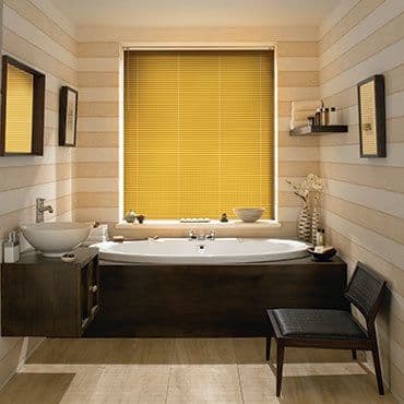 Yellow horizontal blinds fully lowered and closed, thin slats filtering warm light; bathroom with central bathtub, wood-paneled walls, sink, mirrors, shelving, chair and decorative vase.