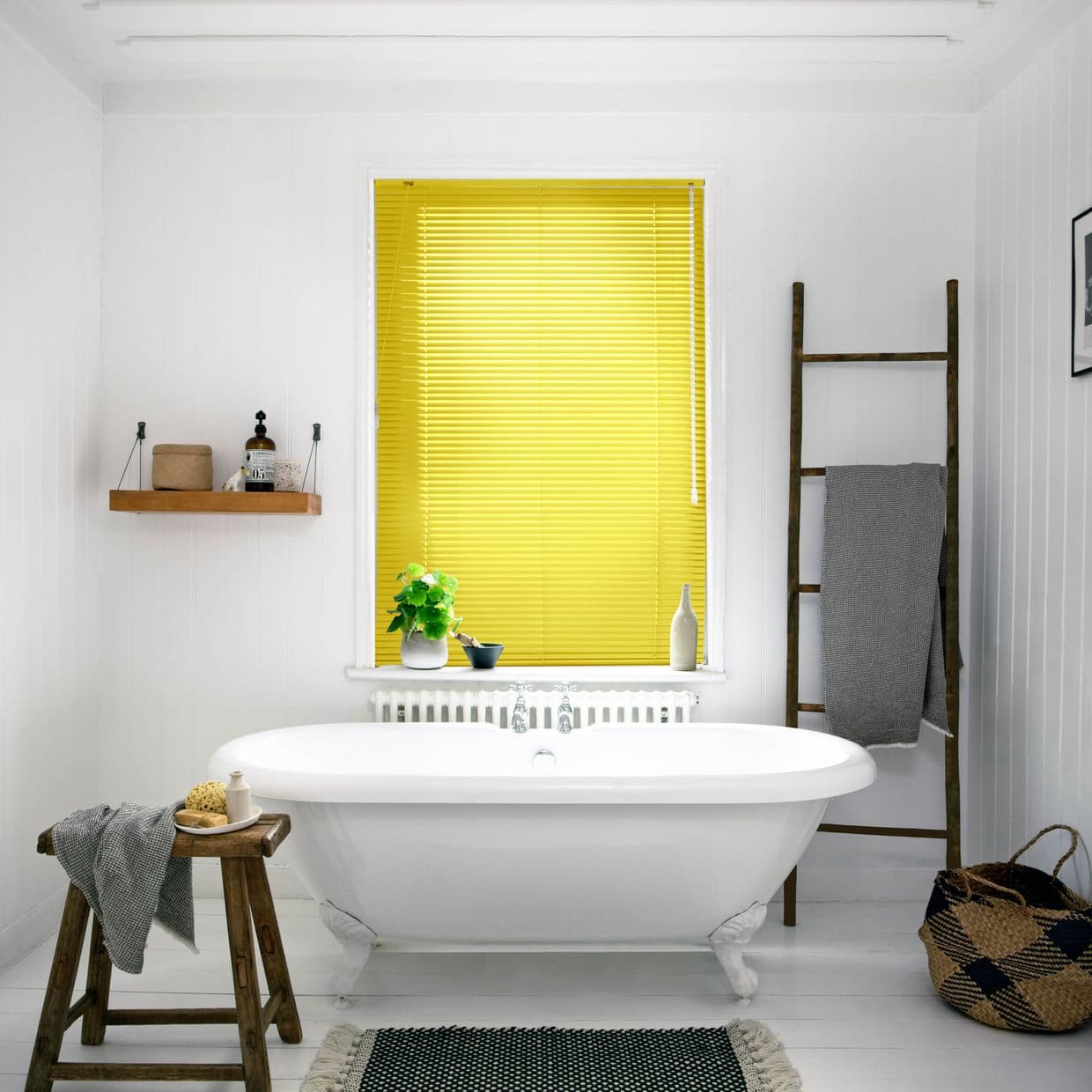 Yellow slatted blinds fully lowered and closed, filtering bright daylight; in a white bathroom with freestanding clawfoot tub, wooden stool, ladder towel rack, woven basket and potted plant.
