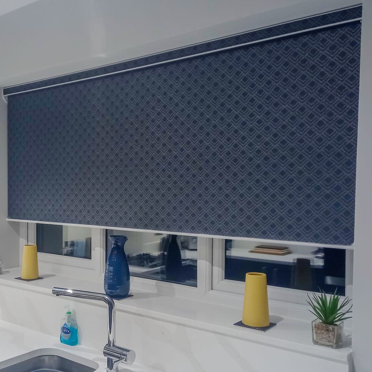 Patterned roller blind (navy diamonds) fully lowered, blocking view and filtering light; kitchen window above sink with faucet, blue vase, yellow votives, soap bottle and small potted plant on sill.

Text in image: Carex