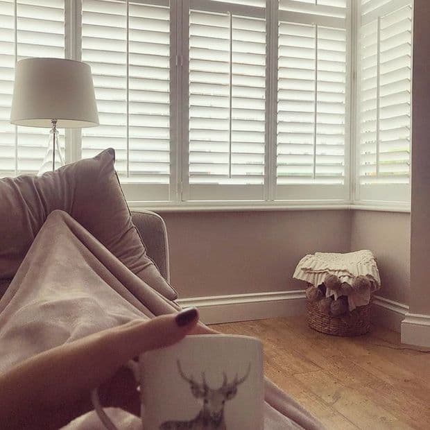 White plantation shutters covering a bay window, closed with angled slats, softly filtering daylight into a cozy living room with sofa, lamp, wicker basket, and foreground hand holding a mug.