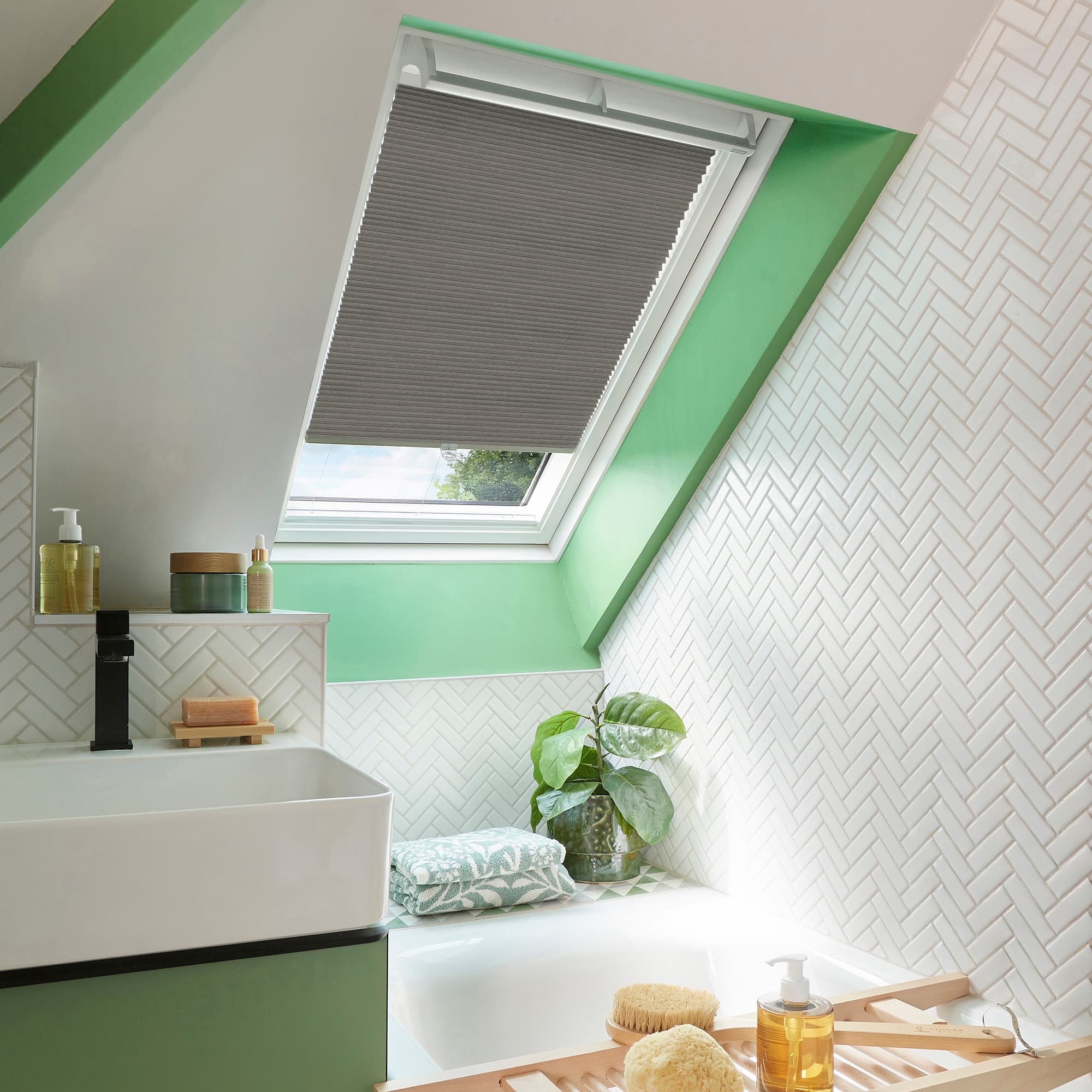 Grey pleated skylight blind lowered to mid-window with a small gap at the bottom, filtering daylight, in bright attic bathroom with green-framed sloped window, white herringbone tiles, sink, tub, plant.