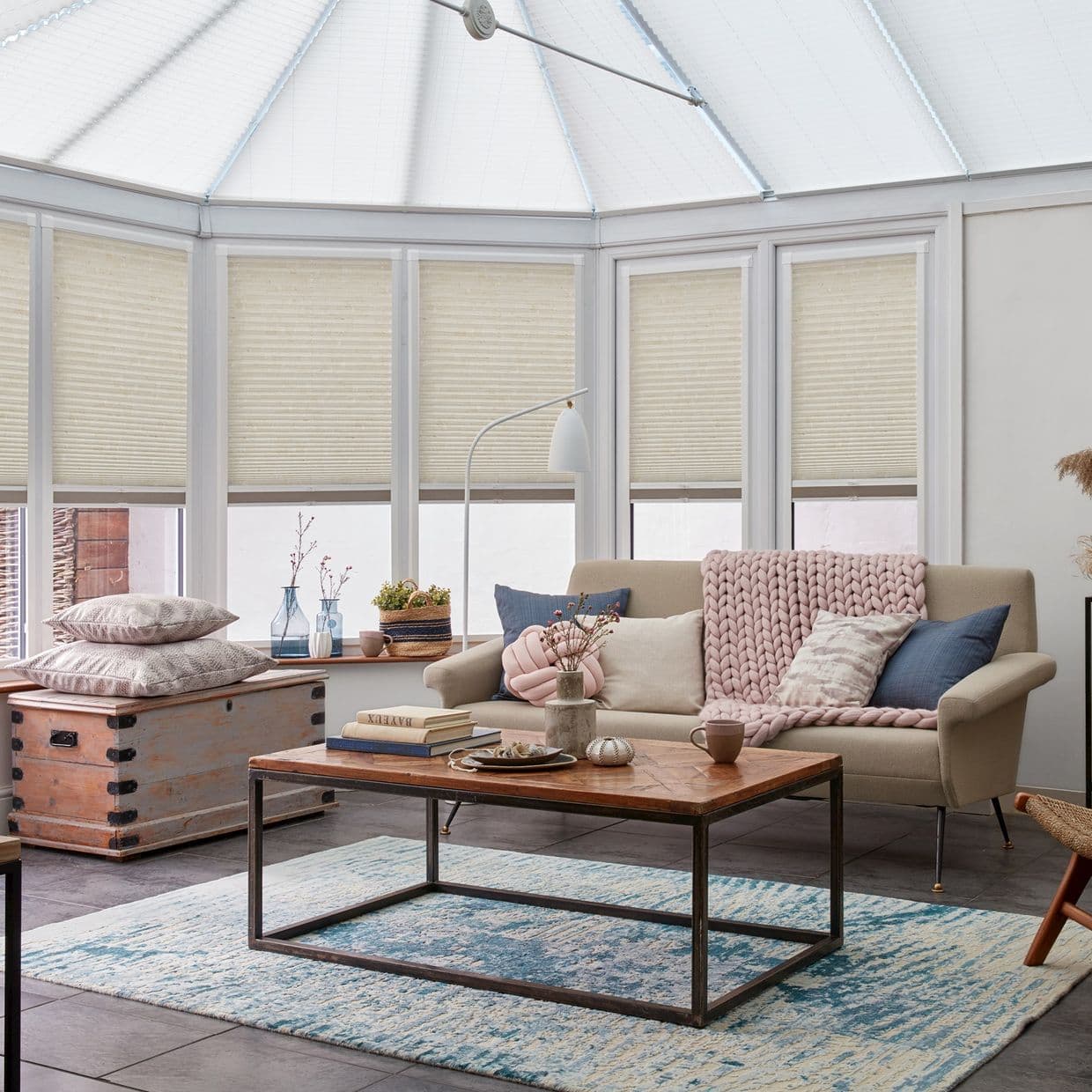 Pleated honeycomb blinds covering conservatoryâ€™s tall, multi-panel windows, lowered to mid-height and filtering soft daylight; beige blinds frame a cozy sunroom with sofa, knitted throw, wooden coffee table and rug.