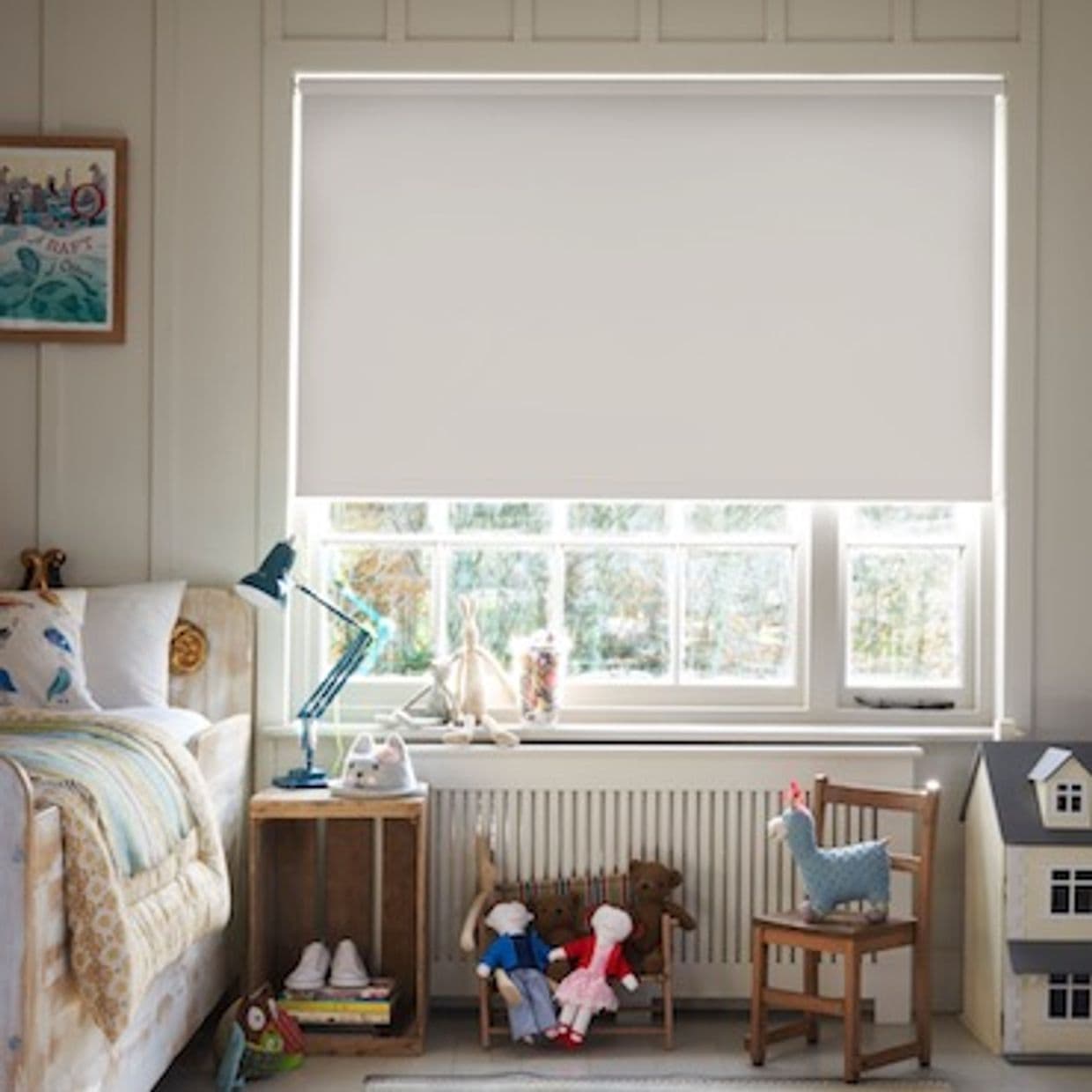 White roller blind lowered over the window, blocking direct view and softly diffusing daylight; in a child's bedroom with a bed, bedside lamp, toys, small chair, radiator and dollhouse beneath.