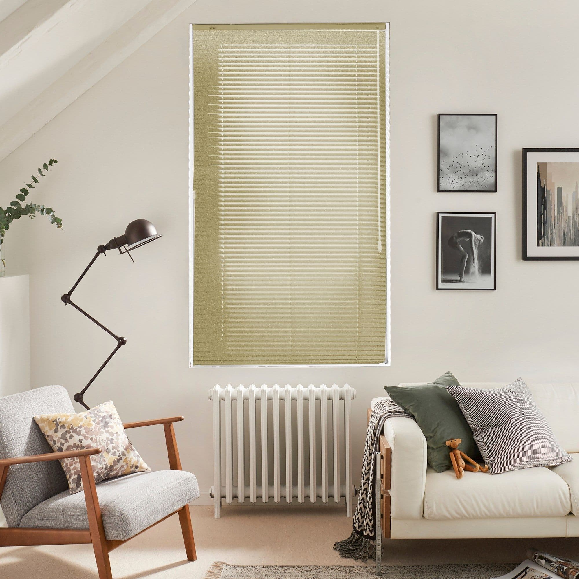 Horizontal beige Venetian blinds covering a tall window, fully closed and softly filtering daylight; living room with armchair, radiator, floor lamp, sofa, cushions and framed wall art in neutral tones.