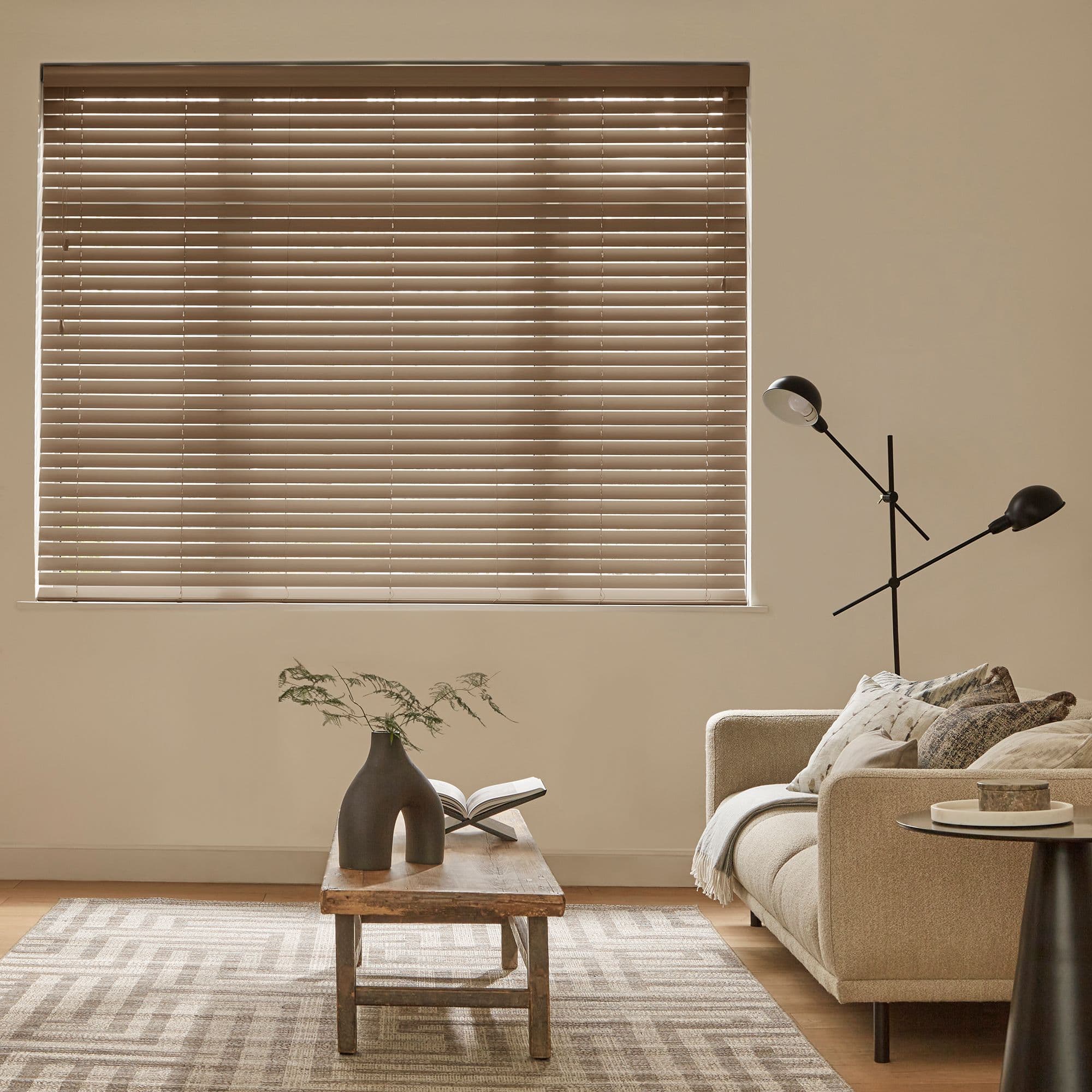 Beige horizontal blinds covering a rectangular window, slatted and mostly closed with thin light slits, filtering daylight into a neutral living room with sofa, wooden coffee table, vase, floor lamp.