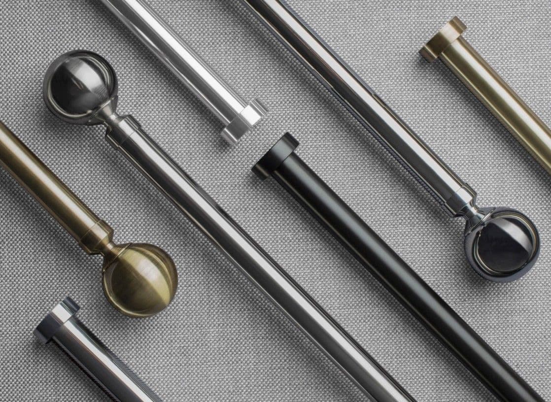Curtain rods with varied metallic finials (spherical, hexagonal), brass, chrome and black finishes, lie diagonally and overlapped, displayed on gray woven fabric under neutral studio lighting.