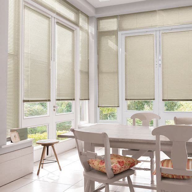 Beige horizontal Venetian blinds covering tall bay windows, lowered and filtering soft daylight; in a bright sunroom dining area with white table, chairs, floral cushions and garden view.